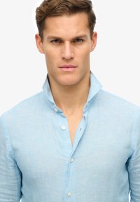 Camicia di lino azzurro chiaro con colletto strutturato, bottoni sul davanti e un aspetto testurizzato. Presenta leggere variazioni di colore.