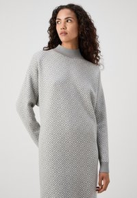 Robe tricotée gris clair avec un col haut, présentant un motif géométrique, des manches longues et des poignets côtelés, offrant une apparence texturée.