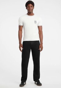 White short-sleeve t-shirt with a circular graphic on the left chest, paired with black straight-leg pants and black sneakers.