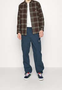 Chemise à carreaux marron avec des accents verts, t-shirt blanc, pantalon cargo bleu avec poches latérales, et baskets navy avec des lacets multicolores.