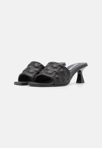 Mules negros de tacón con un upper texturizado de patrón trenzado, diseño de punta abierta y un tacón cilíndrico estrecho.