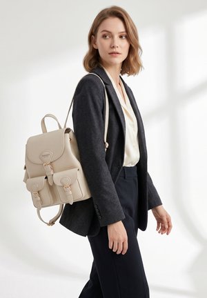 Donna con blazer scuro e camicetta bianca che porta uno zaino in pelle beige, guarda di lato contro uno sfondo chiaro e uniforme.