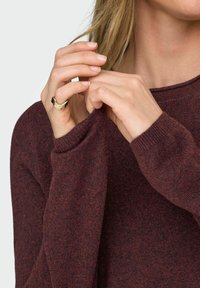 Suéter de punto burdeos con un corte relajado, puños acanalados y un escote amplio. Manos sosteniendo suavemente la manga del suéter, mostrando un anillo de oro.