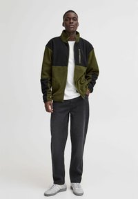 Chaqueta de forro polar con bloques de color negro y verde olivo, cierre de cremallera, cuello alto y bolsillos laterales, combinada con jeans oscuros y zapatillas blancas.
