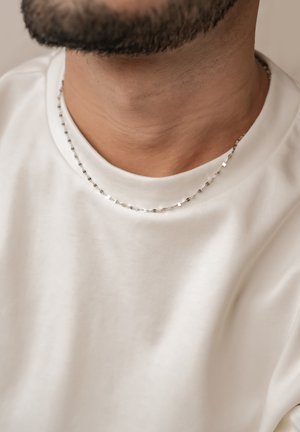 Silver chain necklace with flat, textured links, worn over a plain white crew-neck t-shirt. Close-up focus on neckline and jewelry detail.