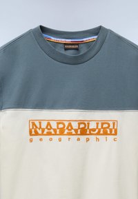 Camiseta de manga corta con un diseño en bloques de color; parte superior en gris oscuro, parte inferior blanca, con un logotipo bordado en naranja "NAPAPIJRI geographic".