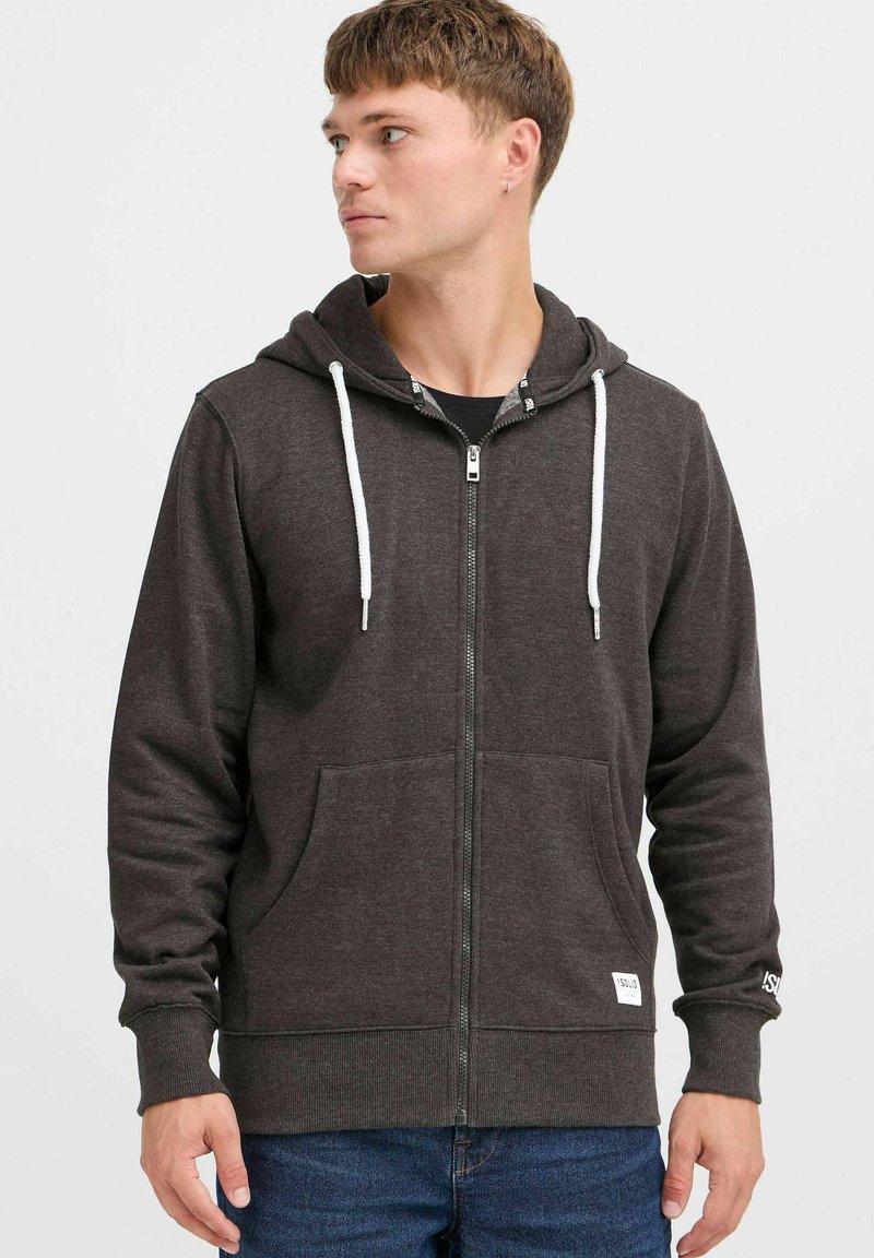 Sweat à capuche gris avec fermeture éclair, doté d'une capuche à cordon de serrage, de poches avant et de poignets côtelés. Il présente une petite étiquette logo au bas et des cordons de serrage blancs contrastants.