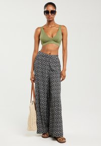 Olīvzaļa, īss krūšturis, melni platmalu bikses ar baltu ziedu rakstu, brūni platformas sandales un krēmkrāsas austs totes soma.