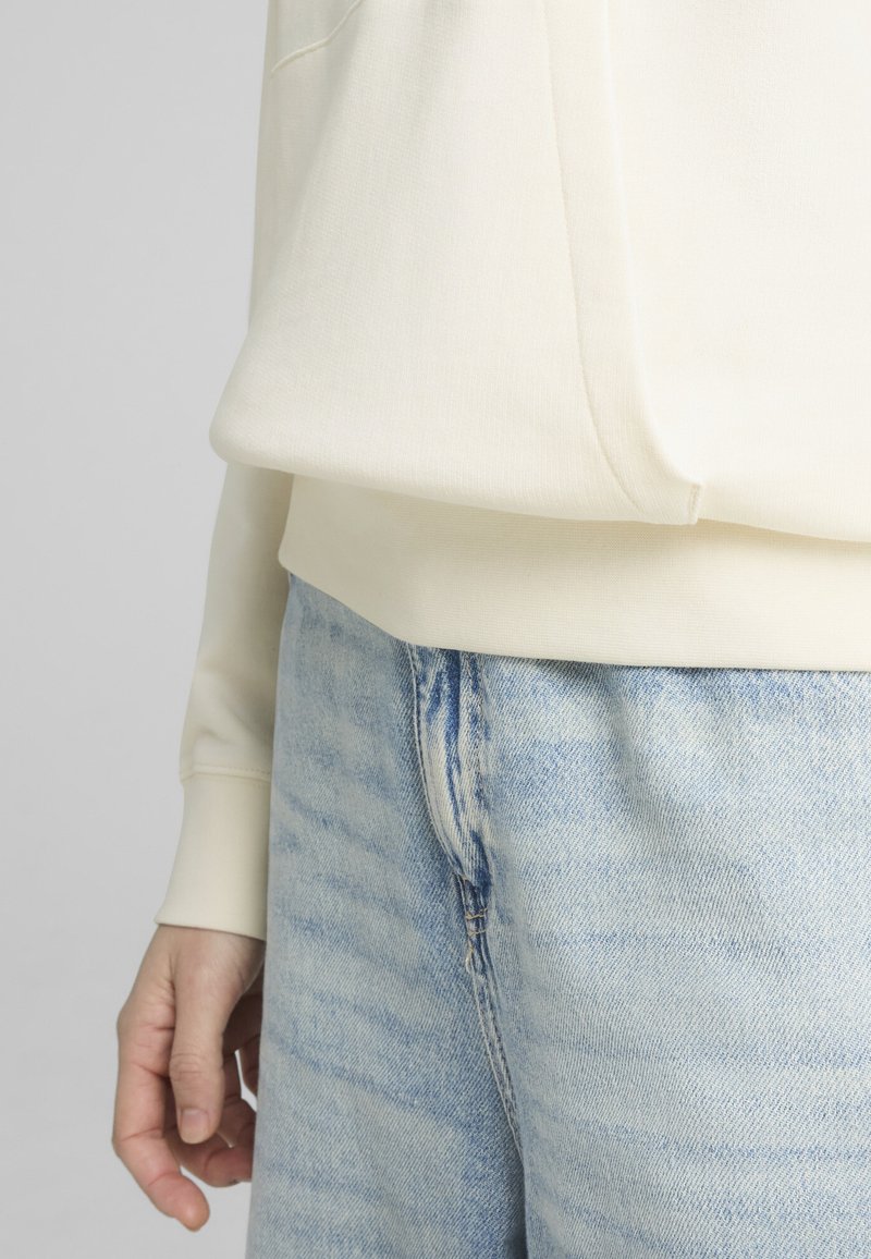 Sudadera color crema con dobladillo cosido, combinada con jeans de lavado claro y desmejorado. Se aprecia su textura suave y ajuste relajado.