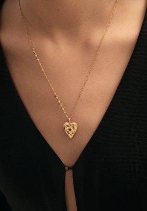 Colgante de oro en forma de corazón con un diseño floral intrincado y una pequeña piedra roja en una cadena fina de oro, llevado en el cuello desnudo de una persona.
