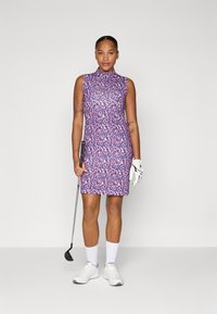 Mugurvējā golfa kleita ar purpursarkanām un rozā leoparda rakstiem, ar augstu apkakli. Uzvilkta ar baltām zeķēm un apaviem, tur rokā golfa nūju.