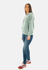 Sudadera con capucha de color verde claro y bolsillo canguro, combinada con jeans azules y zapatillas burdeos con rayas blancas.