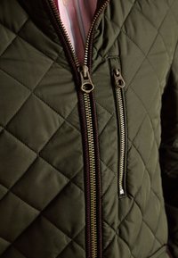 Olivgrüne gesteppt Jacke mit Messing-Reißverschlüssen, ein Hauptreißverschluss teilweise geöffnet, der ein pinkes Hemd darunter zeigt.
