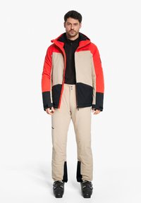 Skijacke mit roten, beigen und schwarzen Einsätzen; verfügt über eine Kapuze, Reißverschlüsse und elastische Bündchen. Beigefarbene Skihose mit schwarzen Akzenten und Knöpfen.