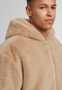Beige-farbene Kunstfell-Hoodiejacke mit einem plüschigen Textur, ausgestattet mit einem Frontreißverschluss und einer lockeren Passform. Betont durch eine große, weiche Kapuze.