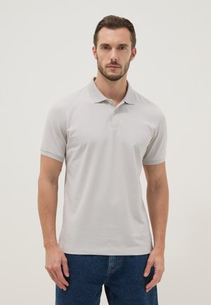 Mann mit kurzen dunklen Haaren, der ein hellgraues Poloshirt und blaue Jeans trägt, vor einem einfarbigen hellen Hintergrund stehend.