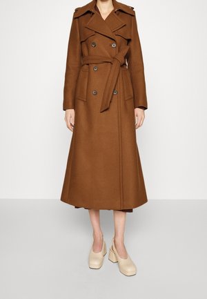 Brauner, doppelreihiger, langer Wollmantel mit weitem Kragen, gebundener Taille, Klappentaschen, getragen mit beigen, blockabsatzgeschlossenen Schuhen.