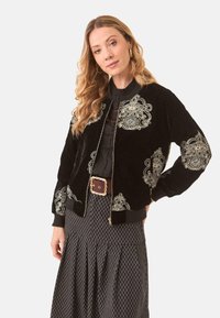 Cazadora bomber de terciopelo negro con bordado floral dorado en las mangas y el frontal; puños y bajo de canalé; cierre de cremallera dorado.
