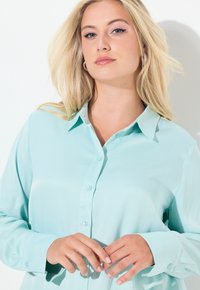 Lyseblå, langærmet skjorte med knapper og krave, glat tekstur og matchende knapper. Model poserer foran en hvid baggrund.