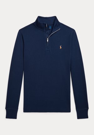 Tamsiai mėlynas ilgomis rankovėmis puloveris su pusiau užtrauktuku ant kaklo, su raštuotomis rankogaliais ir maža siuvinėta emblema kairėje krūtinės pusėje.