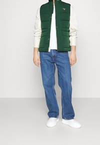 Muž v zelené prošívané vestě, béžové košili s dlouhým rukávem, modrých džínách a bílých teniskách stojící proti jednobarevnému pozadí.