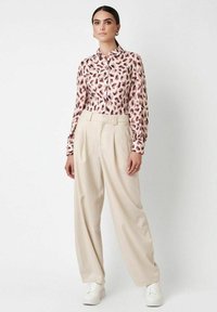 Frau trägt eine gemusterte Bluse mit langen Ärmeln, hochgeschnittene beige weit geschnittene Hose und weiße Sneakers, stehend vor einem schlichten Hintergrund.