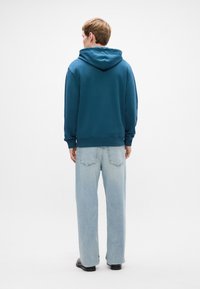 Mėlynas džemperis su gobtuvu, su elastingomis rankovėmis ir apačia. Dėvimas su šviesiai mėlynomis plačiomis džinsomis. Matoma nugaros dalis be logotipų.