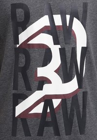 Szara koszulka z wyraźnym nadrukiem w czerni i bieli, z dużym czerwonym akcentem, prezentująca warstwowy design słów "RAW 3 RAW."