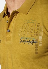 Yellow polo shirt with a textured finish, featuring a buttoned placket, embroidered graphics, and contrasting blue accents.