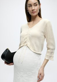 Ανοιχτό μπεζ πλεκτό πουλόβερ με V-λαιμόκοψη και κλείσιμο με κορδόνι, με μανίκια 3/4 και σχέδιο που μοιάζει με δαντέλα. Φορεμένο πάνω από μια λευκή μίντι φούστα με υφή.