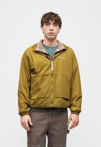 Νεαρός άνδρας που φοράει μπουφάν Columbia μουσταρδί με μπροστινές τσέπες, στέκεται μπροστά σε ένα απλό ανοιχτό γκρι φόντο.