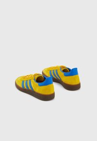 Zapatillas de ante amarillas con rayas azules, parte superior texturizada, suela de goma marrón y punta redonda. Presenta cordones planos y talón reforzado.