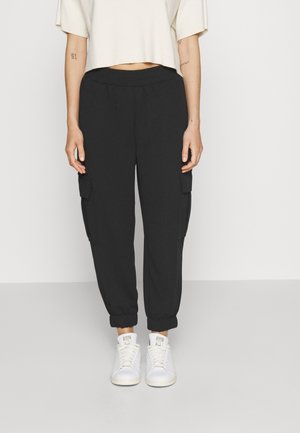 Personne portant un pantalon cargo jogger noir avec des poignets élastiques et des baskets blanches, debout devant un fond clair uni.