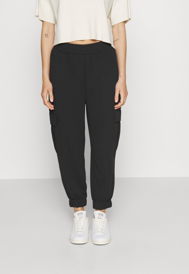 Personne portant un pantalon cargo jogger noir avec des poignets élastiques et des baskets blanches, debout devant un fond clair uni.