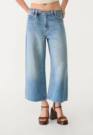 Kvinde iført lyseblå vide cropped jeans og brune højhælede sandaler, stående mod en ensfarvet hvid baggrund.