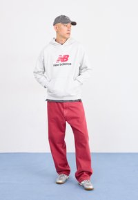 Junger Mann trägt einen grauen New Balance Hoodie, rote Hose, silberne Sneaker und eine graue Mütze. Er steht auf einem blauen Boden vor einer weißen Wand.