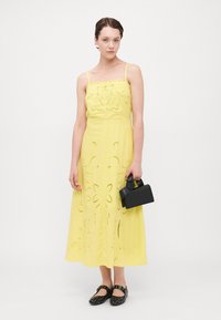 Vestido maxi amarillo sin mangas con patrones de flores recortados, textura suave y silueta acampanada, acompañado de un pequeño bolso negro.