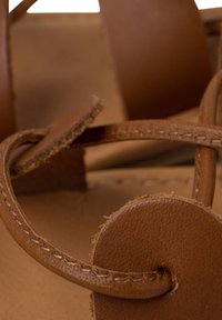 Sandal in pelle marrone con fascette incrociate, accentuazione rotonda e dettagli a bordo grezzo. Superfici texturizzate e lisce con cuciture.