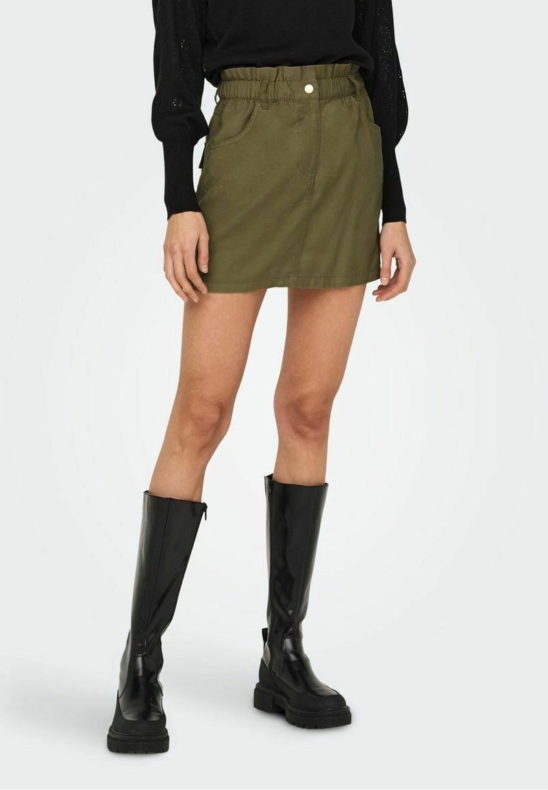 Femme portant une mini-jupe vert olive avec ceinture élastique et poches avant, un haut noir à manches longues et des bottes noires montantes jusqu'aux genoux.