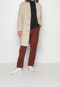 Persona che indossa un cappotto beige texture sopra una maglietta nera, pantaloni color ruggine e sneakers bianche, in piedi con una mano in tasca.