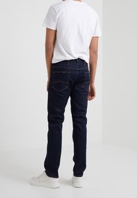 En person står vänd bort med en vit t-shirt, mörkblå jeans med synliga bakfickor och vita sneakers på en neutral bakgrund.