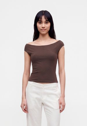 Kvinde med langt mørkt hår iført en brun off-shoulder top og hvide bukser, står foran en ensfarvet lys baggrund.