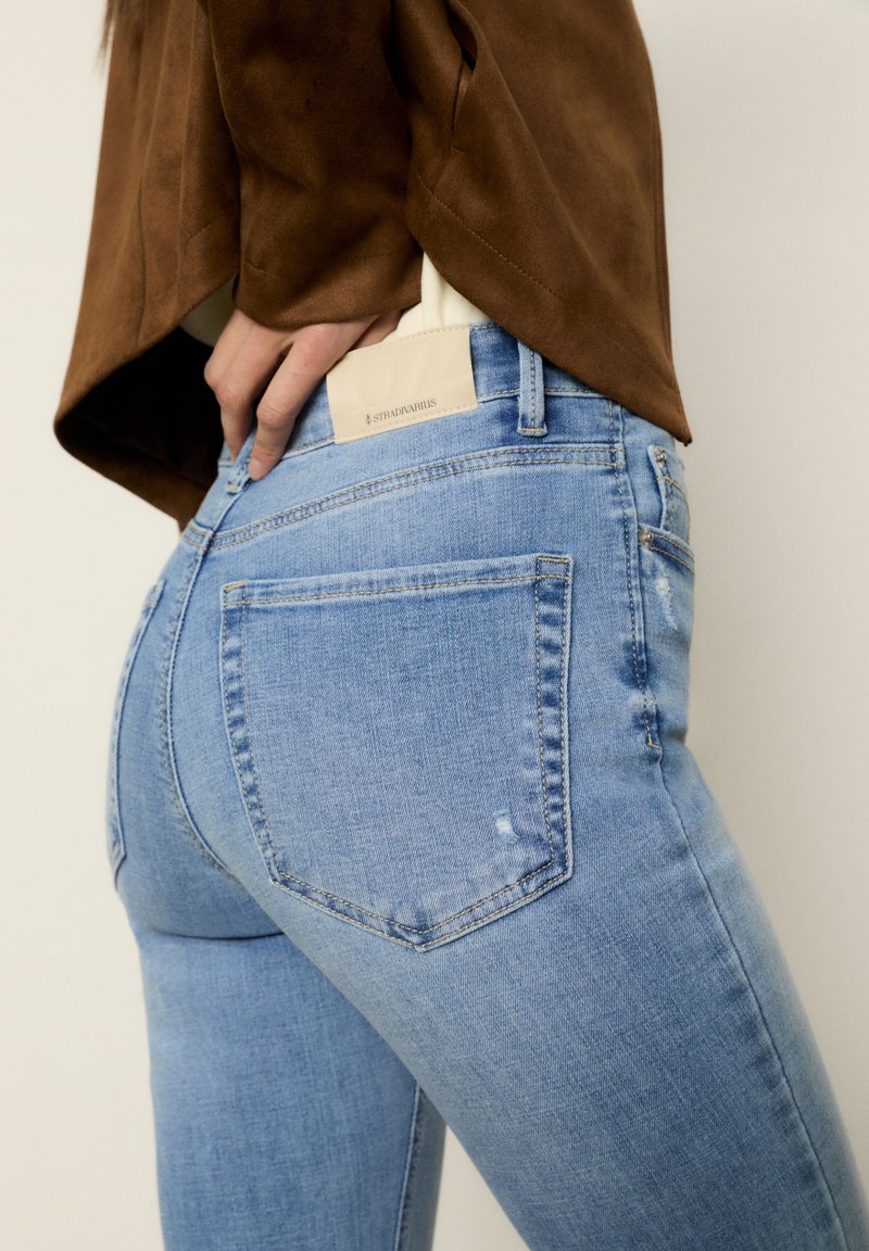 Personne portant un jean bleu clair ajusté avec une poche arrière et un haut marron, main reposant près de la taille.
