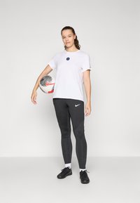 White short-sleeve shirt with a round navy emblem, paired with black patterned leggings and black athletic shoes, holding a soccer ball.