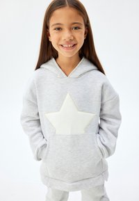 Sudadera gris claro con un gran parche en forma de estrella blanca en el frente, un bolsillo frontal y un corte holgado con capucha ajustable.