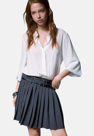 Giovane donna con capelli mossi che indossa una camicetta bianca larga con bottoni e una gonna plissettata grigio scuro con doppie cinture con fibbia.