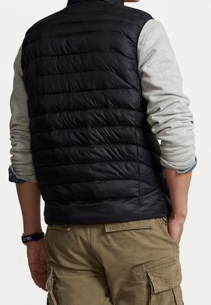 Mann trägt eine schwarze gesteppte Weste über einem grauen Langarmshirt, khakifarbene Cargohosen und ein schwarzes Armband, Hände in den Taschen, von hinten gesehen.