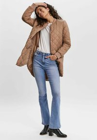 Quilted beige long jacket with ribbed collar, paired with light blue flared jeans and black ankle boots. Smooth texture and casual fit.