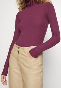 Langsleeve coltrui van diep paarse geribbelde stof met aansluitend ontwerp. Gecombineerd met hooggetailleerde beige broek met voorrits en zakken.