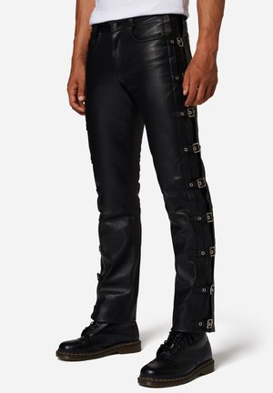 Schwarze Lederhosen mit schlanker Passform, seitlichen Schnallenakzenten und Kontrastnähten. Kombiniert mit schwarzen Schnürstiefeln.